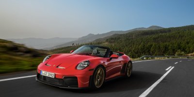 A red Porsche 911 GT3 RS convertible drives along a winding mountain road under a clear blue sky. The car is in motion, with blurred wheels and a scenic backdrop of forested hills and distant mountains. The driver is visible in the open cockpit.